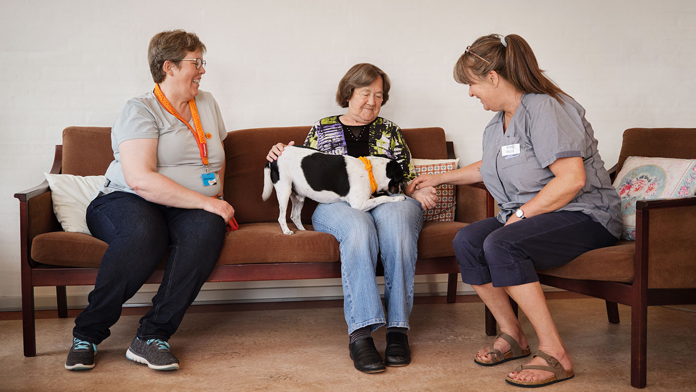 En beboer sidder i en sofa med besøgshunden Uffe i skødet sammen med ejer Henriette og en medarbejder.